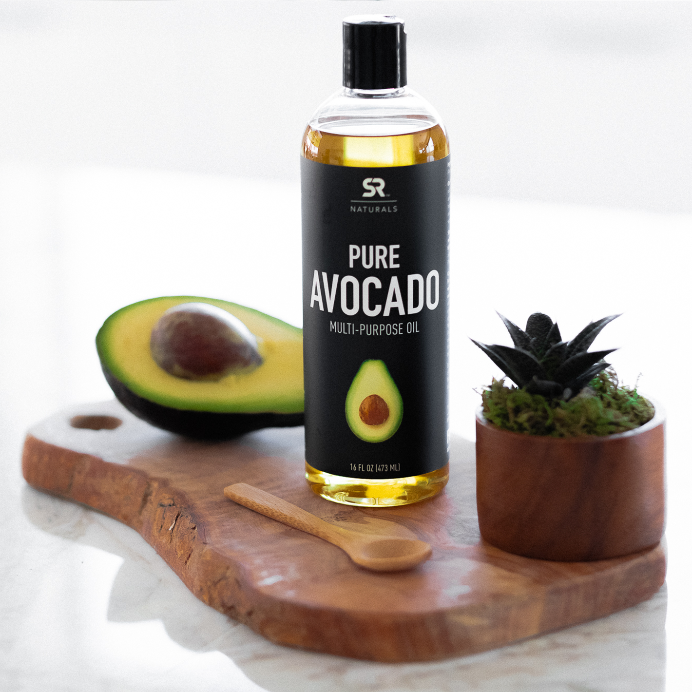 A bottle of Sports Research Naturals Pure Avocado Oil sitting next to a half sliced avocado and a small plant on a wooden board.