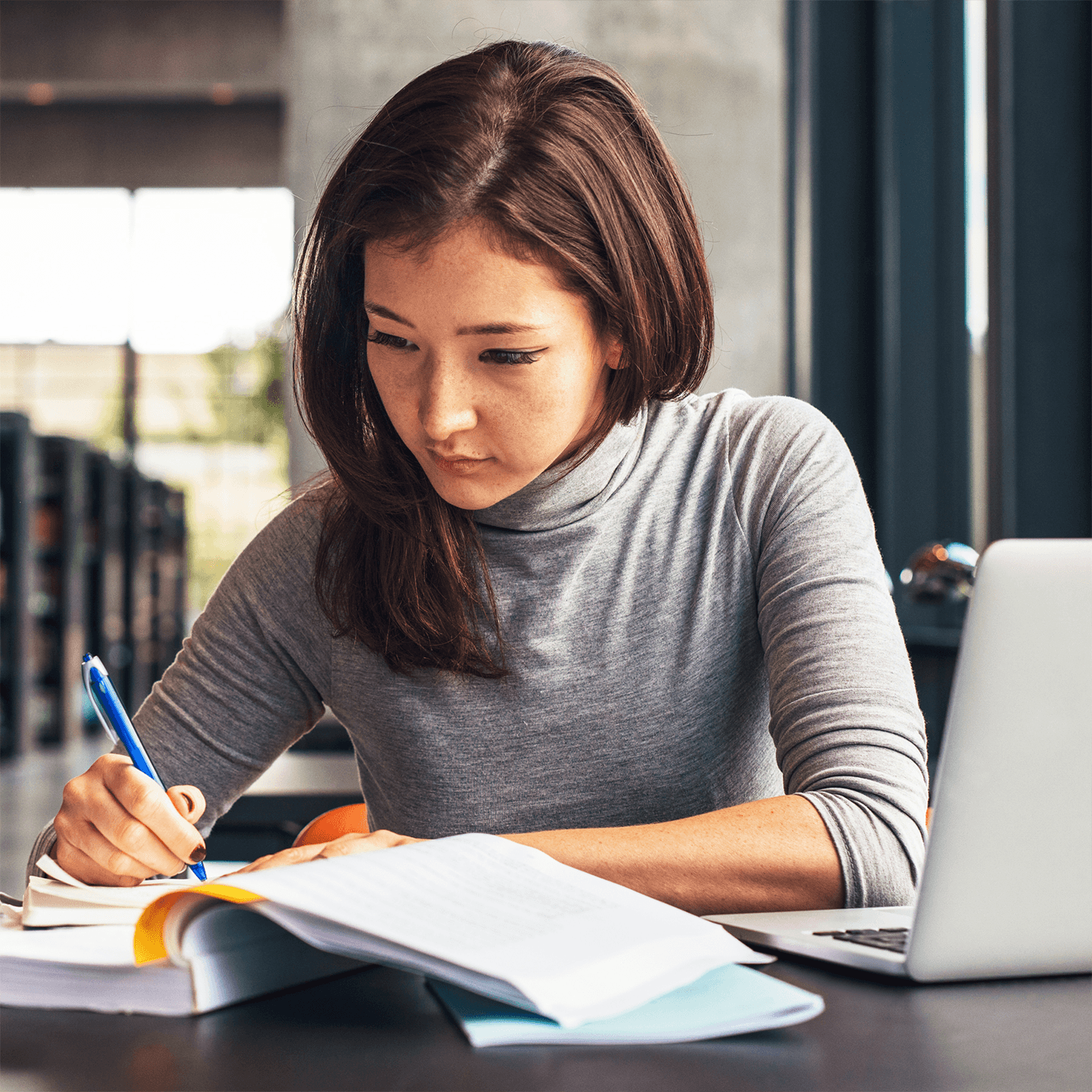 Woman in a library studying with a textbook and laptop computer in front of her.