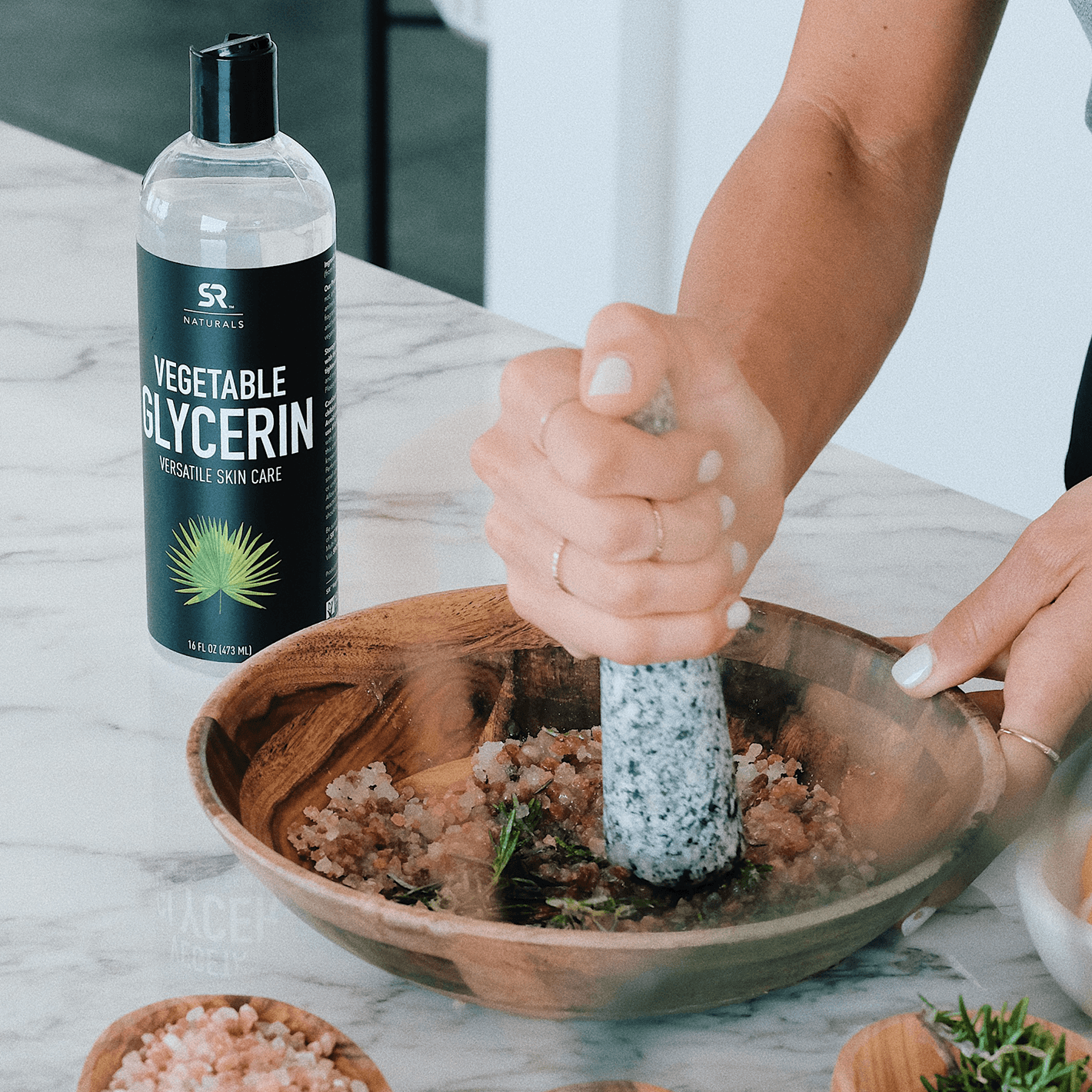 A woman grinding vegetables in a wooden bowl with a bottle of Sports Research Naturals Vegetable Glycerin on the counter next to it.