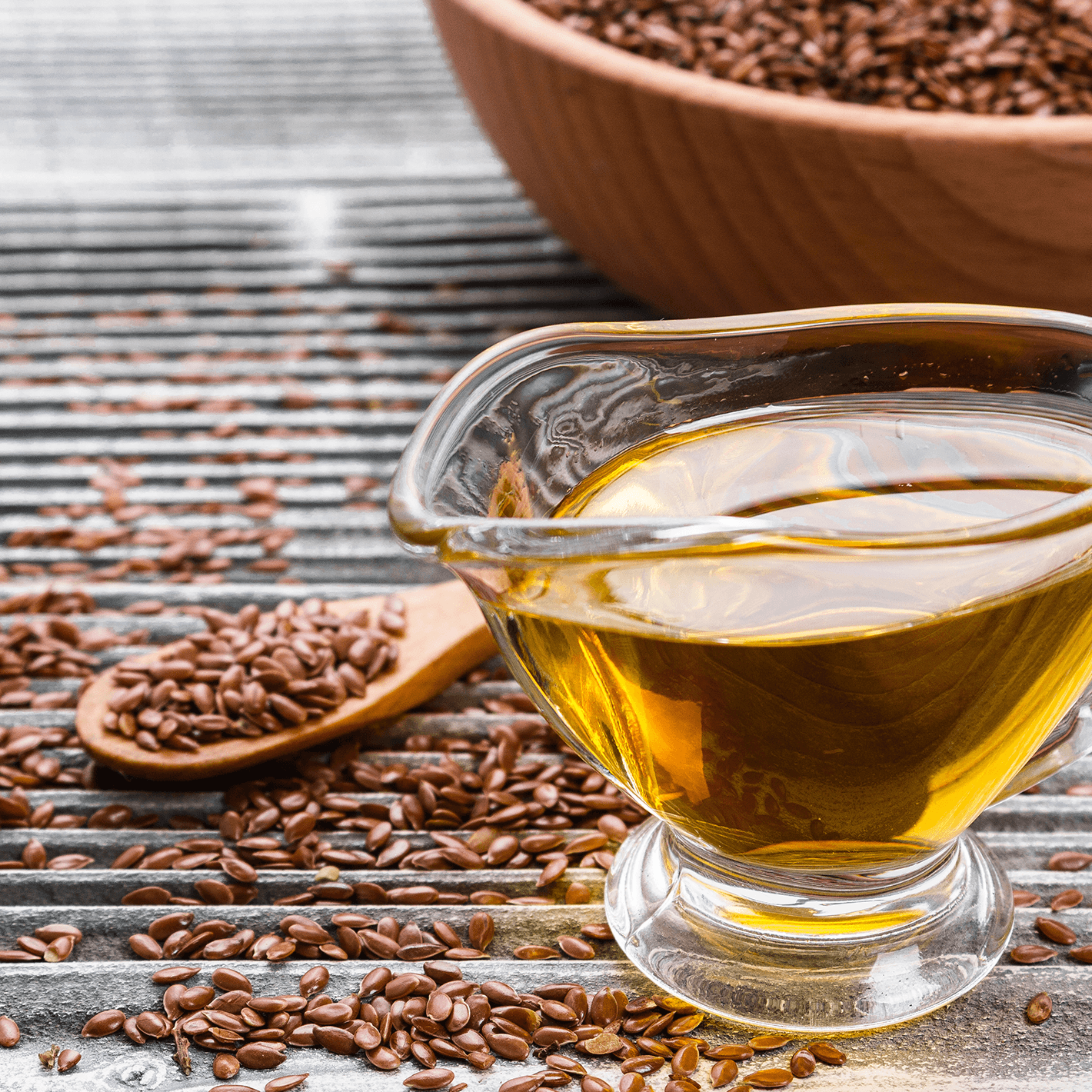 A small glass gravy boat with oil in it with a bowl of flaxseed behind and scattered around.