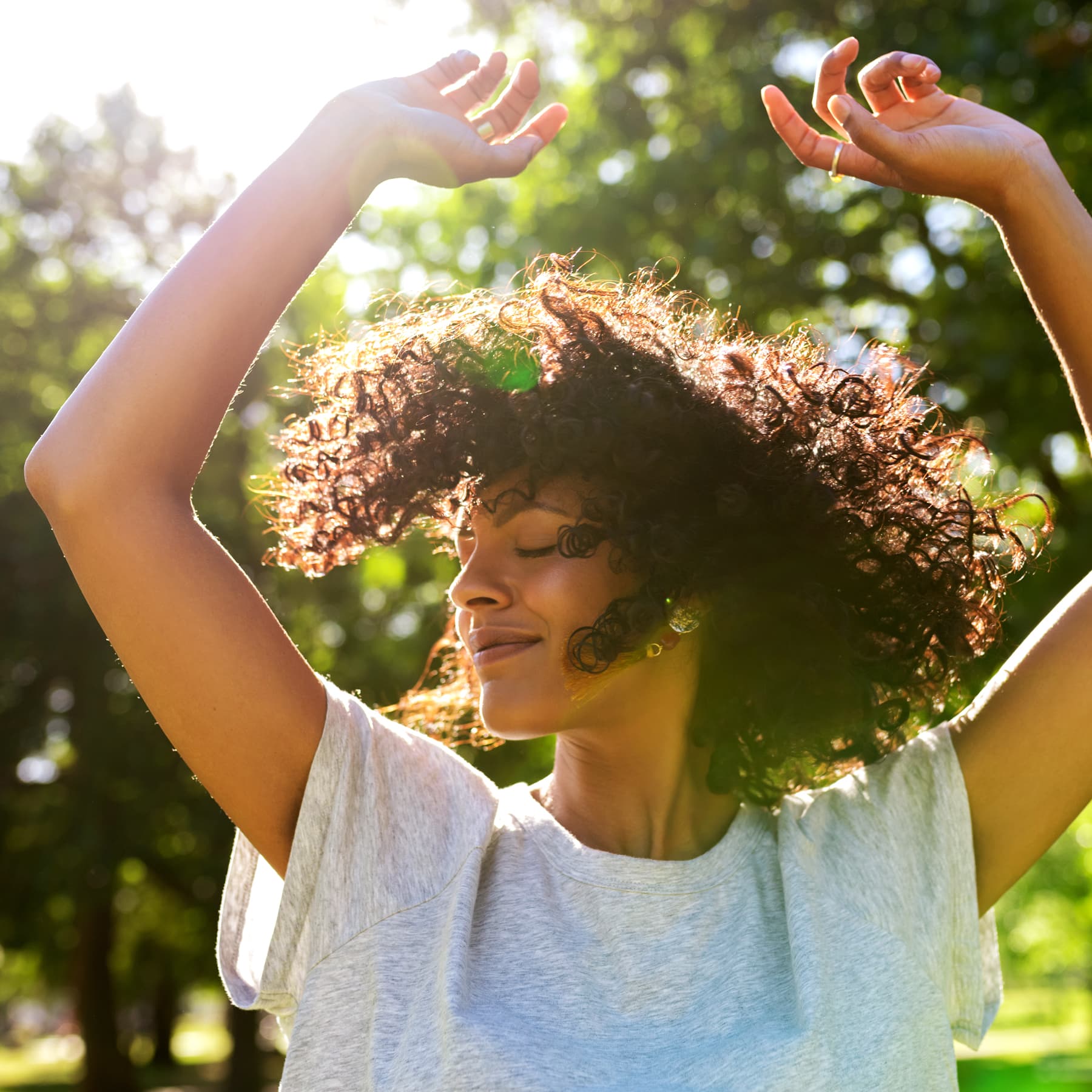 Woman outdoors standing in the sunshine with arms stretched upwards.