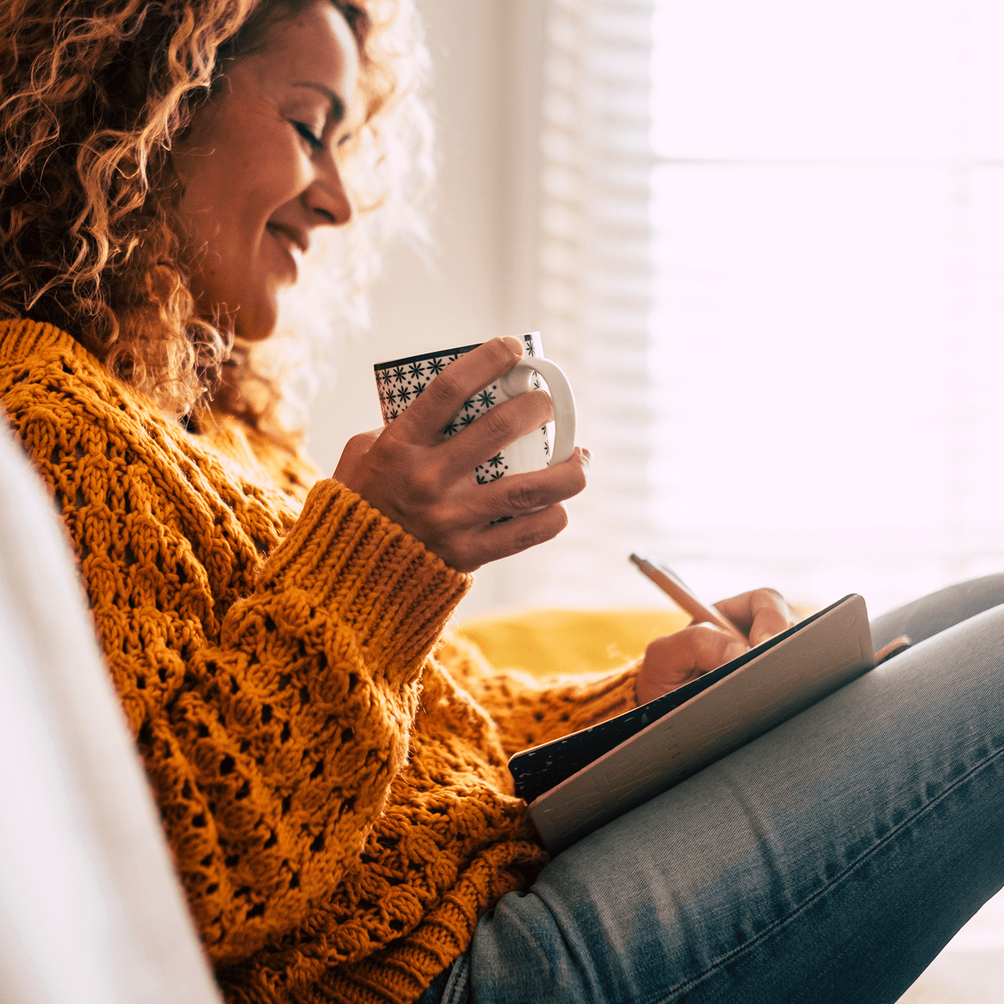 Woman sitting on a couch writing in a journal, while holding a coffee cup.